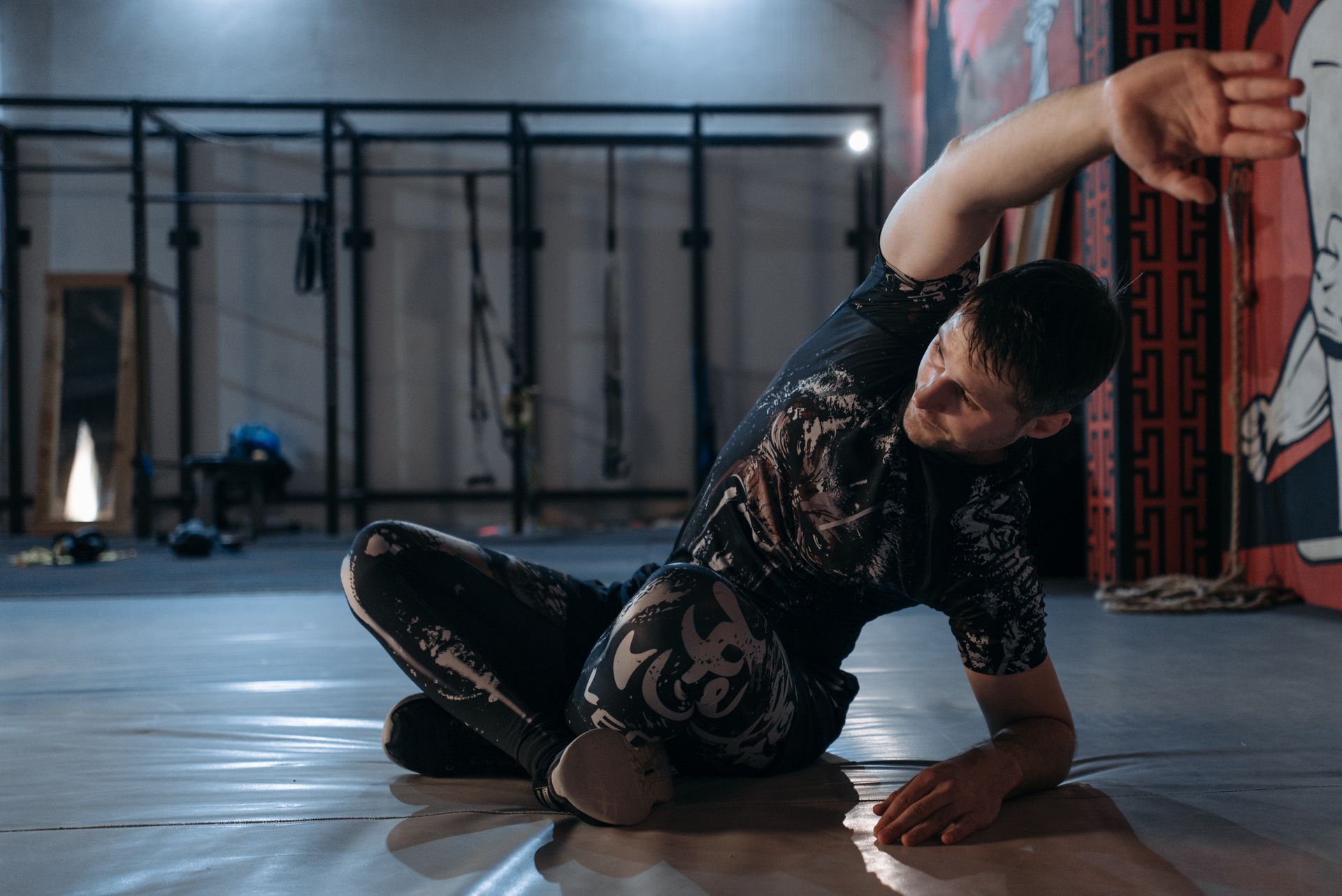 A man performs a stretching routine indoors in a mixed martial arts gym, preparing for training.