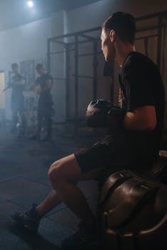 A boxer sits thoughtfully in a gym, wearing black gloves and attire, surrounded by muted lighting.