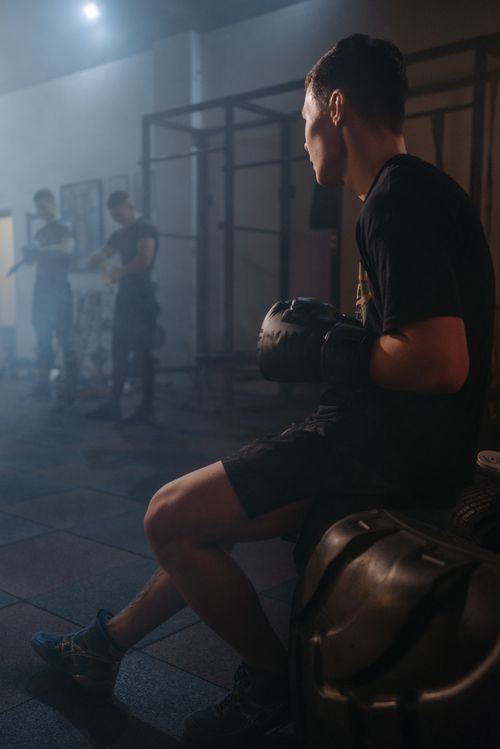 Free A boxer sits thoughtfully in a gym, wearing black gloves and attire, surrounded by muted lighting. Stock Photo