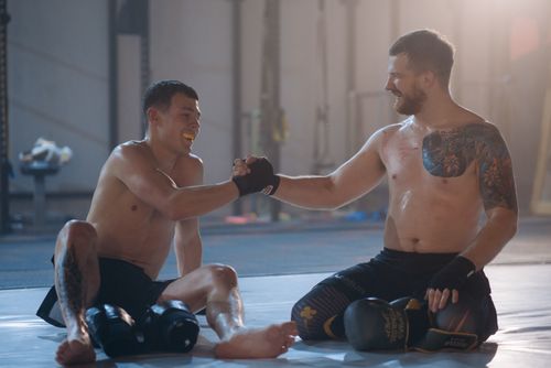 Free Two male martial artists sitting on the mat post-training, embracing sportsmanship with a handshake. Stock Photo