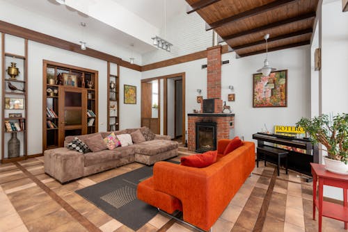 Colorful sofas placed on parquet in room with decorative elements on shelves and brick fireplace near piano and green plant in apartment