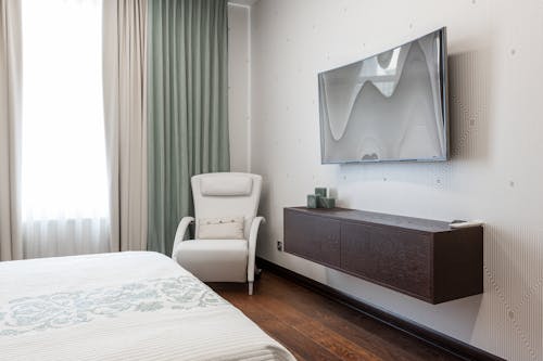 Interior of modern bedroom with white blanket on bed and armchair placed near window at wall with modern TV set