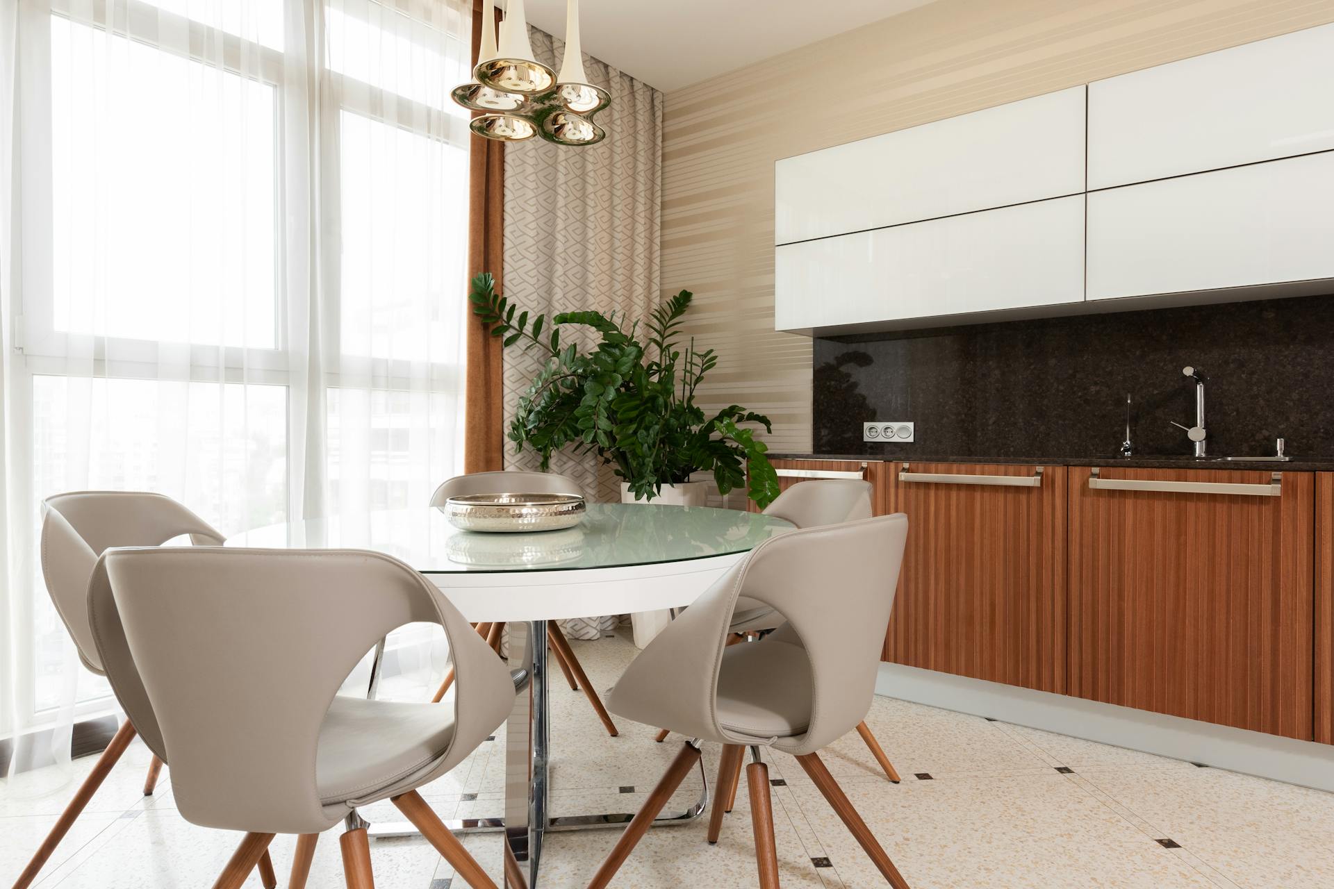 Plate on white round table with stools placed on tiled floor near green potted plant and counter with sink and wooden cupboards in dining room