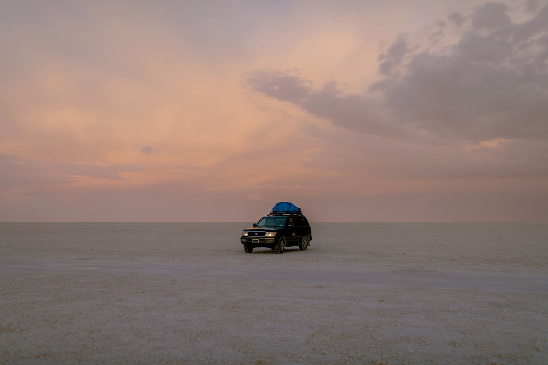 Lonely black automobile driving through empty sandy desert against cloudy sky in sunset