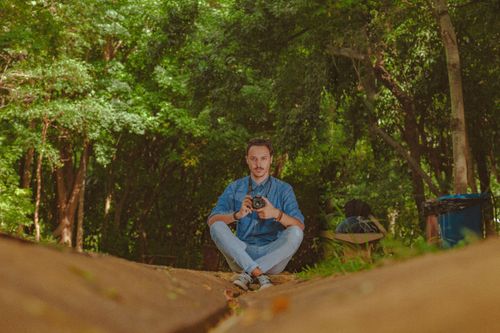 Free Ground level full body of male photographer with crossed legs sitting on ground among green plants and looking at camera Stock Photo