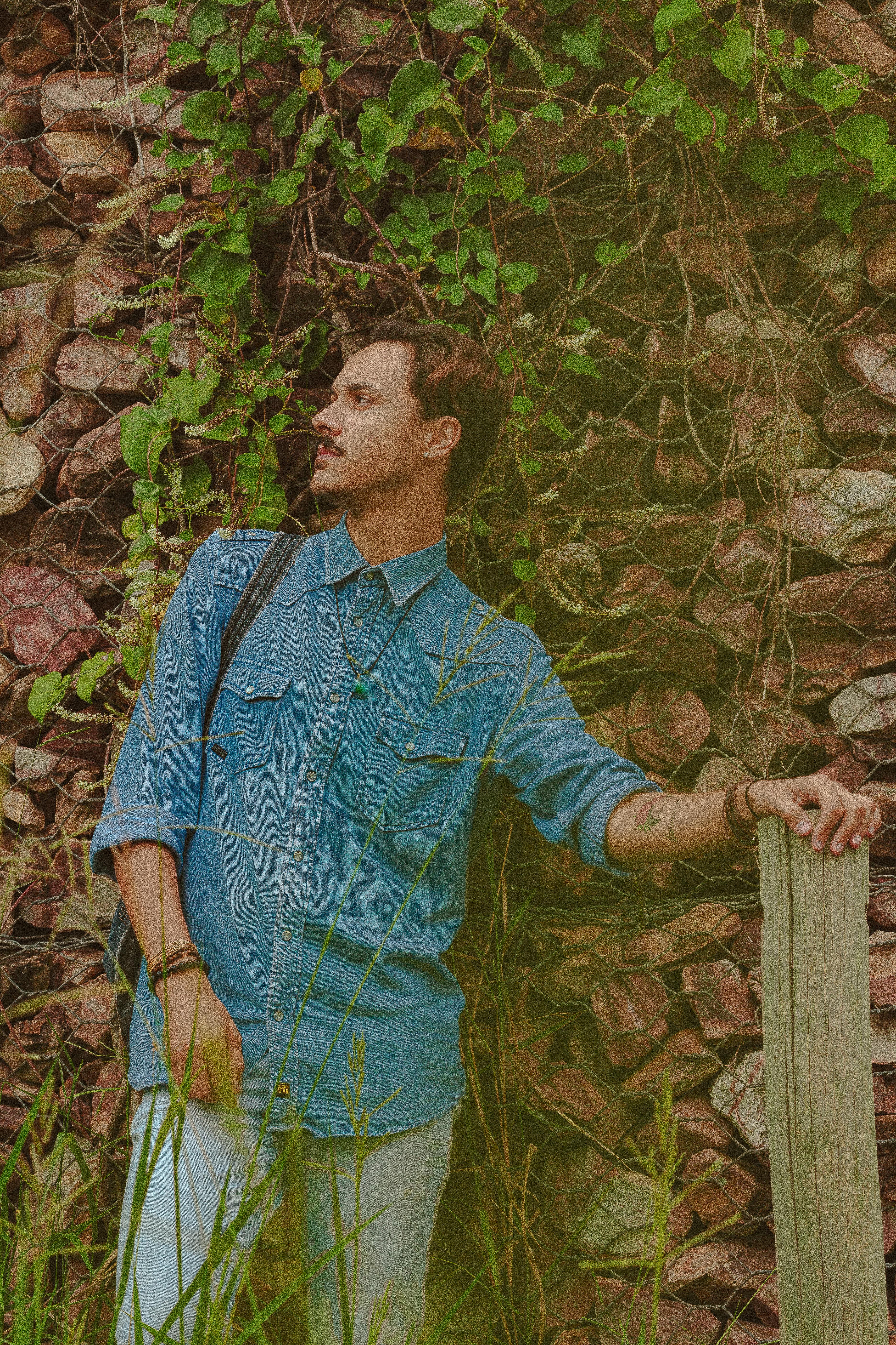 Calm male with mustache wearing denim shirt looking into distance while standing near wall with rocks in countryside