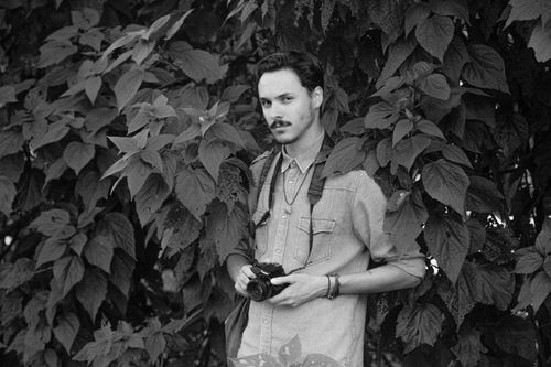 Free Black and white of serious male photographer with photo camera looking at camera while standing near tree branches in park Stock Photo