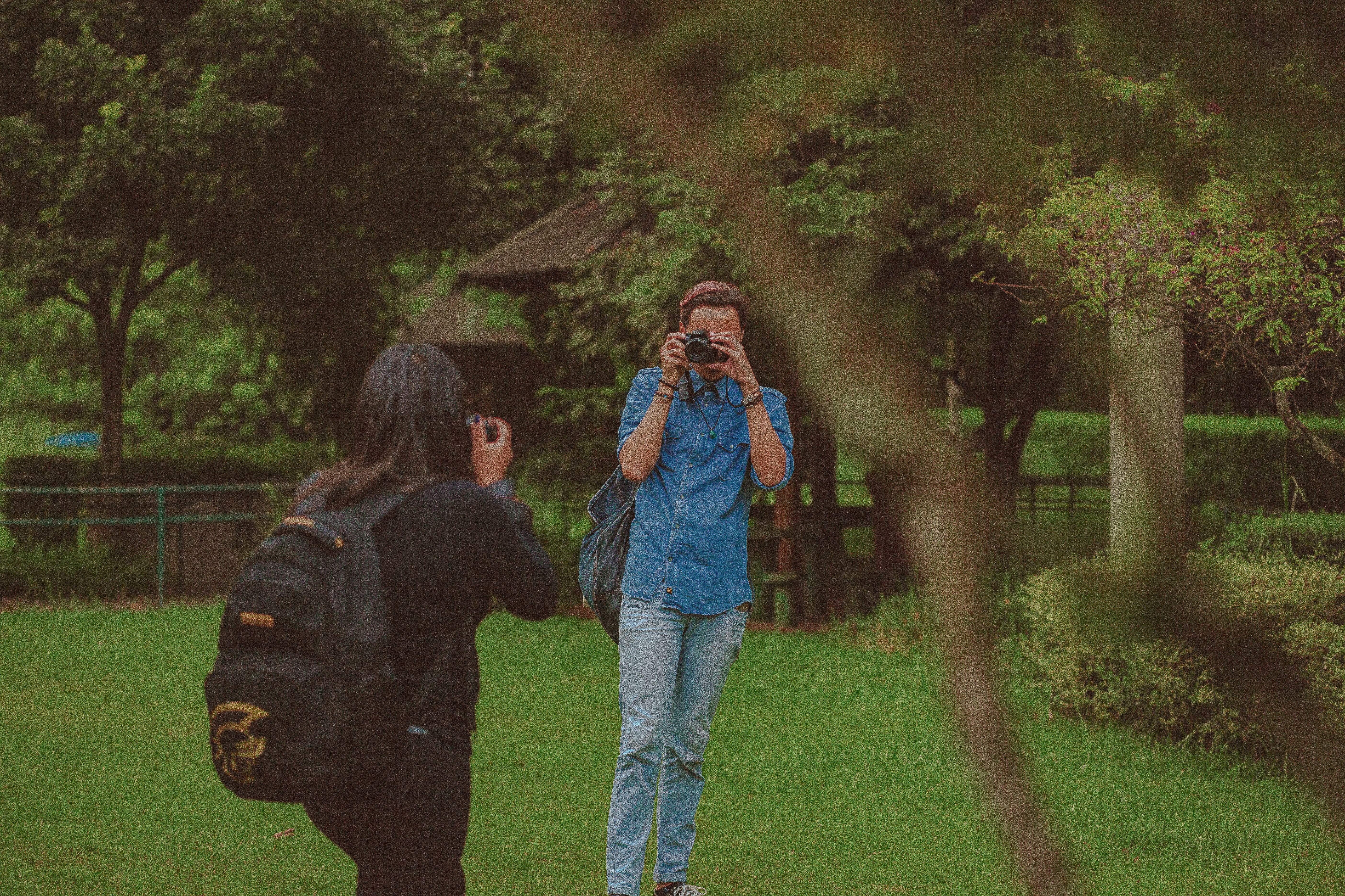 Anonymous man and woman with photo cameras taking pictures of each other while standing on grassy lawn in park with green trees