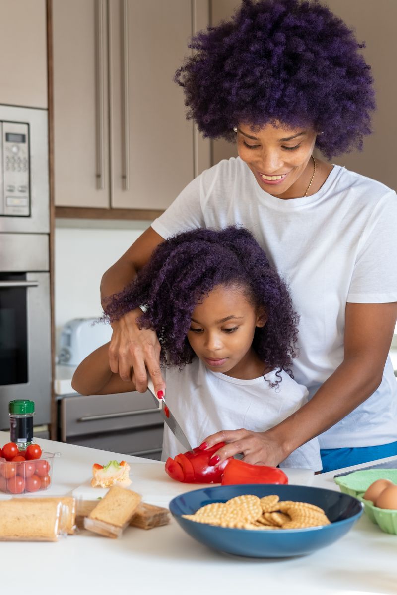 A Black mother and her daughter cook together at a kitchen counter, slicing a fresh red bell pepper among vegetables, crackers, and eggs.