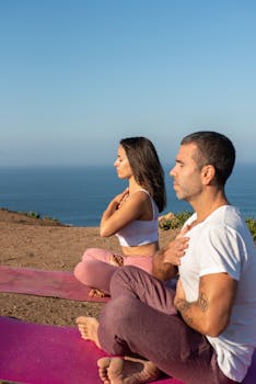 A couple practicing meditation on yoga mats by the ocean, focusing on mindfulness and relaxation.