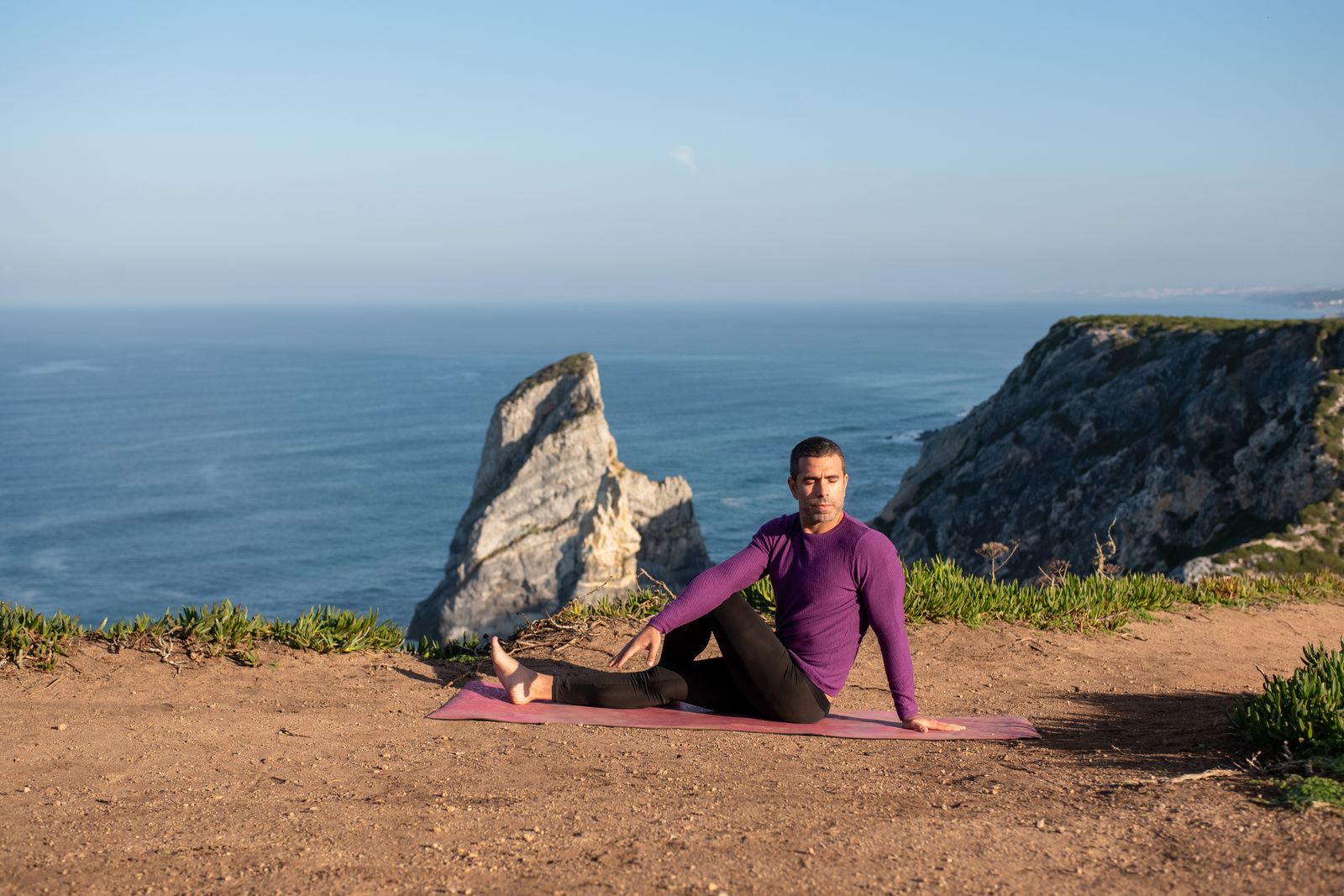 Person practicing a seated yoga spinal twist pose outdoors near cliffs and ocean
