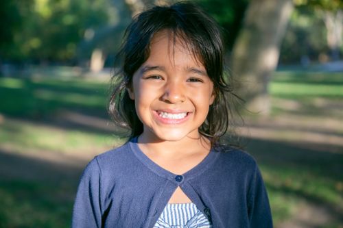 Free A young girl smiling brightly outdoors in a sunlit park, capturing joyful childhood moments. Stock Photo