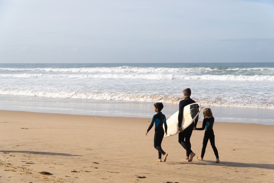 A father and two children walk with a surfboard on a sunny Portuguese beach.