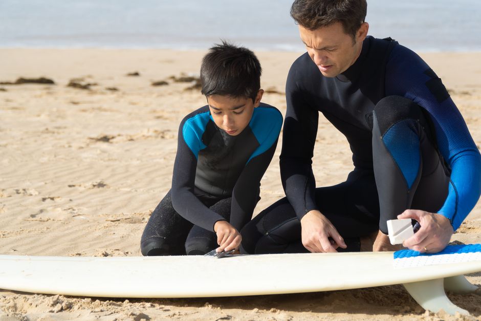 A father and son wearing wetsuits prepare a surfboard on a sandy beach in Portugal.