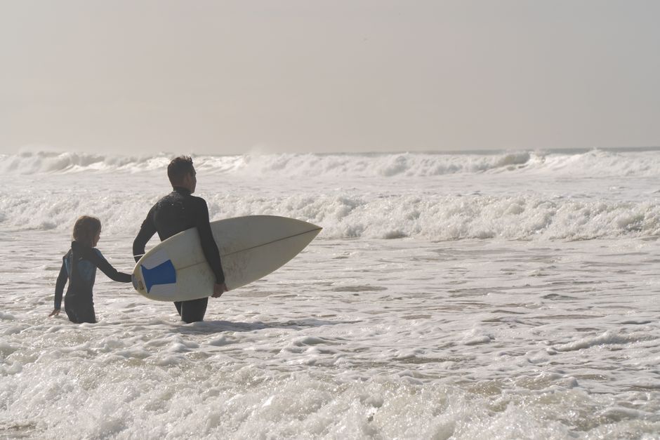 A father and child wade through ocean waves with a surfboard, enjoying a sunny day at the beach in Portugal.