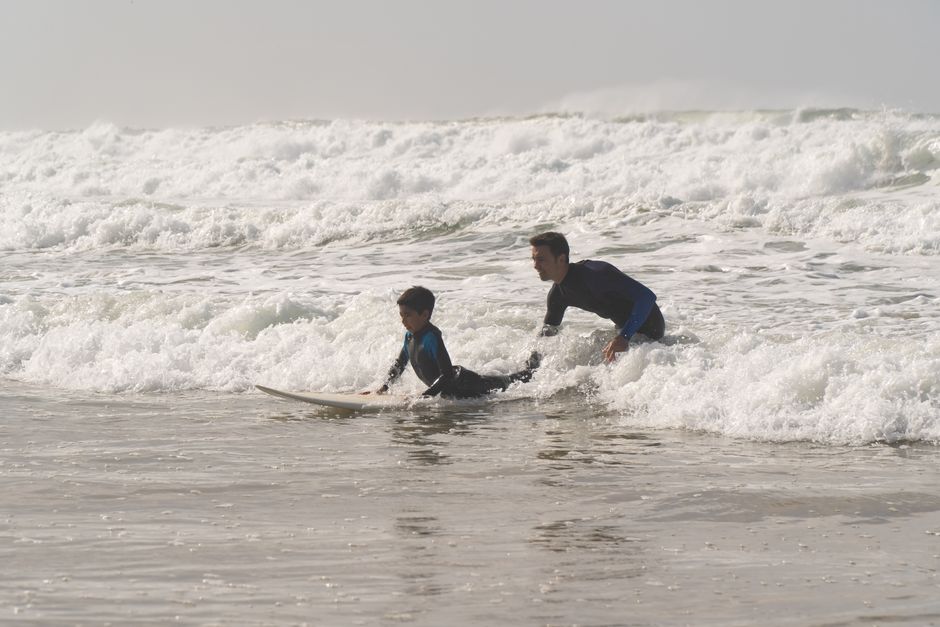 A dad guides his child on a surfboard at a lively Portuguese beach, teaching surfing basics.