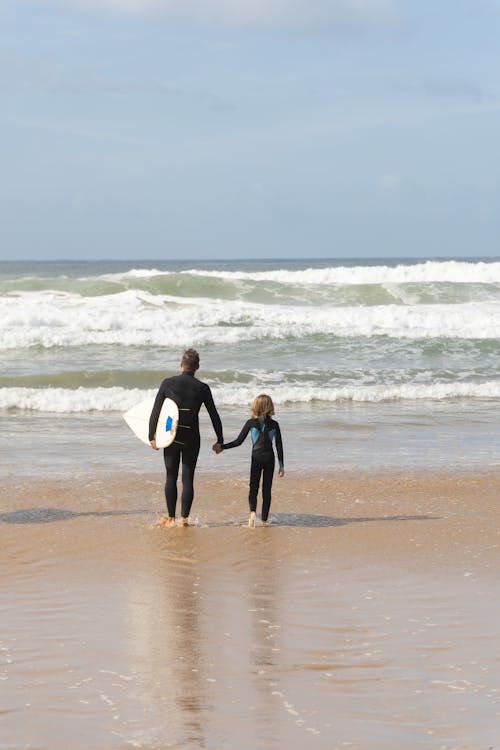 A Man Carrying a Surfboard and a Child Walking on Beach Shore