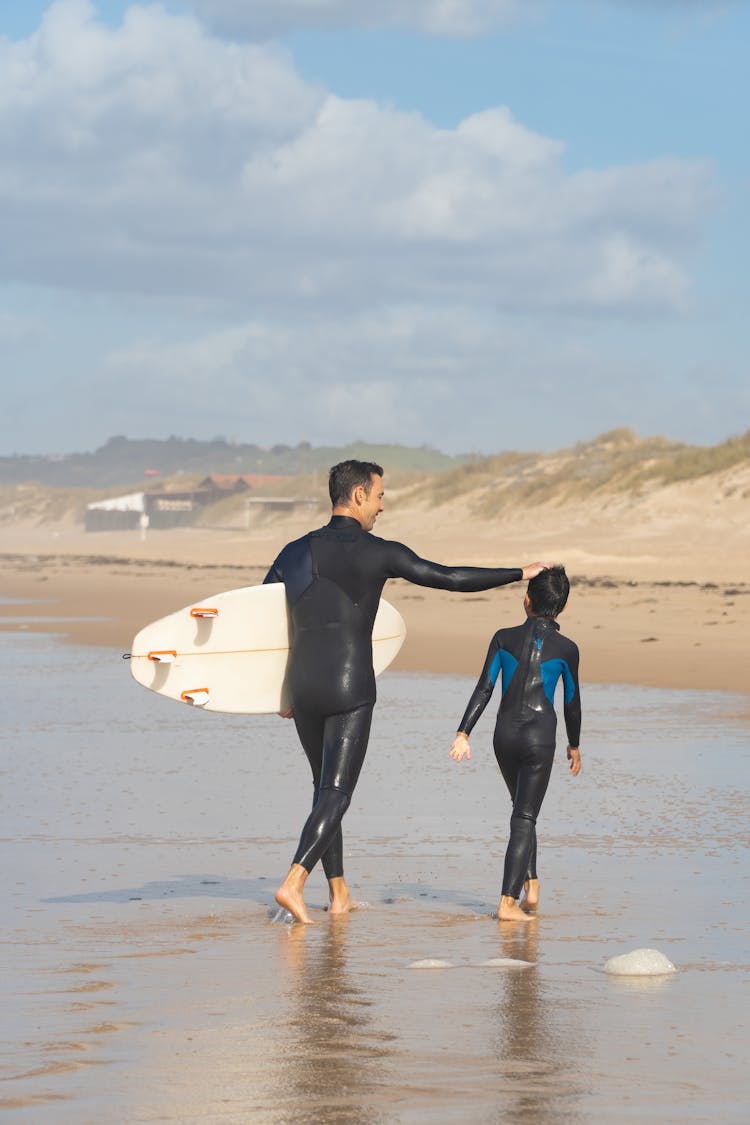 A Man And A Boy In Wetsuits Walking On Beach Shore