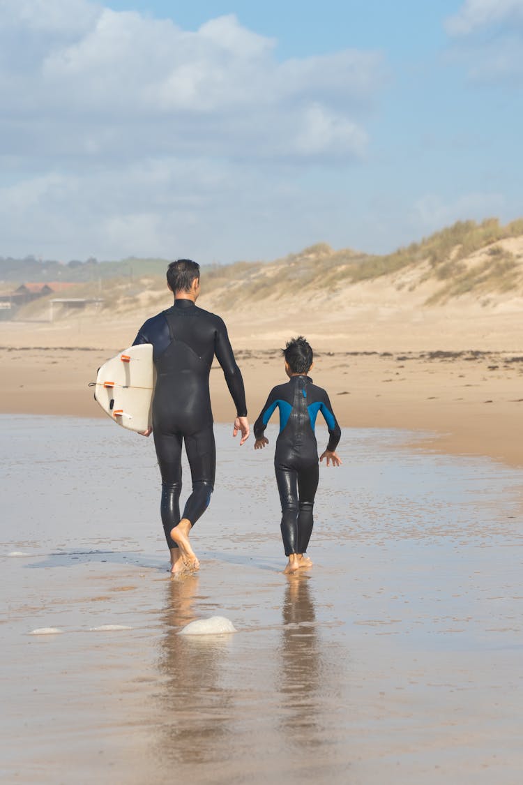 Boy And A Man In Wetsuits Carrying A Surfboard