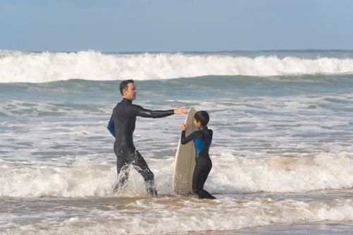 Free Father teaching son to surf on a sunny beach in Portugal with waves crashing in the background. Stock Photo