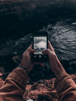 Hands capturing a rugged Moroccan coastline with a smartphone from above.