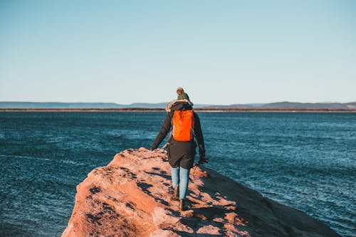 Free Back view of anonymous female explorer in warm outerwear and hat walking on rocky cliff with photo camera in hand and admiring sea on sunny day Stock Photo