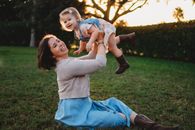 Side view happy young female in casual wear raising adorable content daughter on hands while sitting on lush grass in verdant park