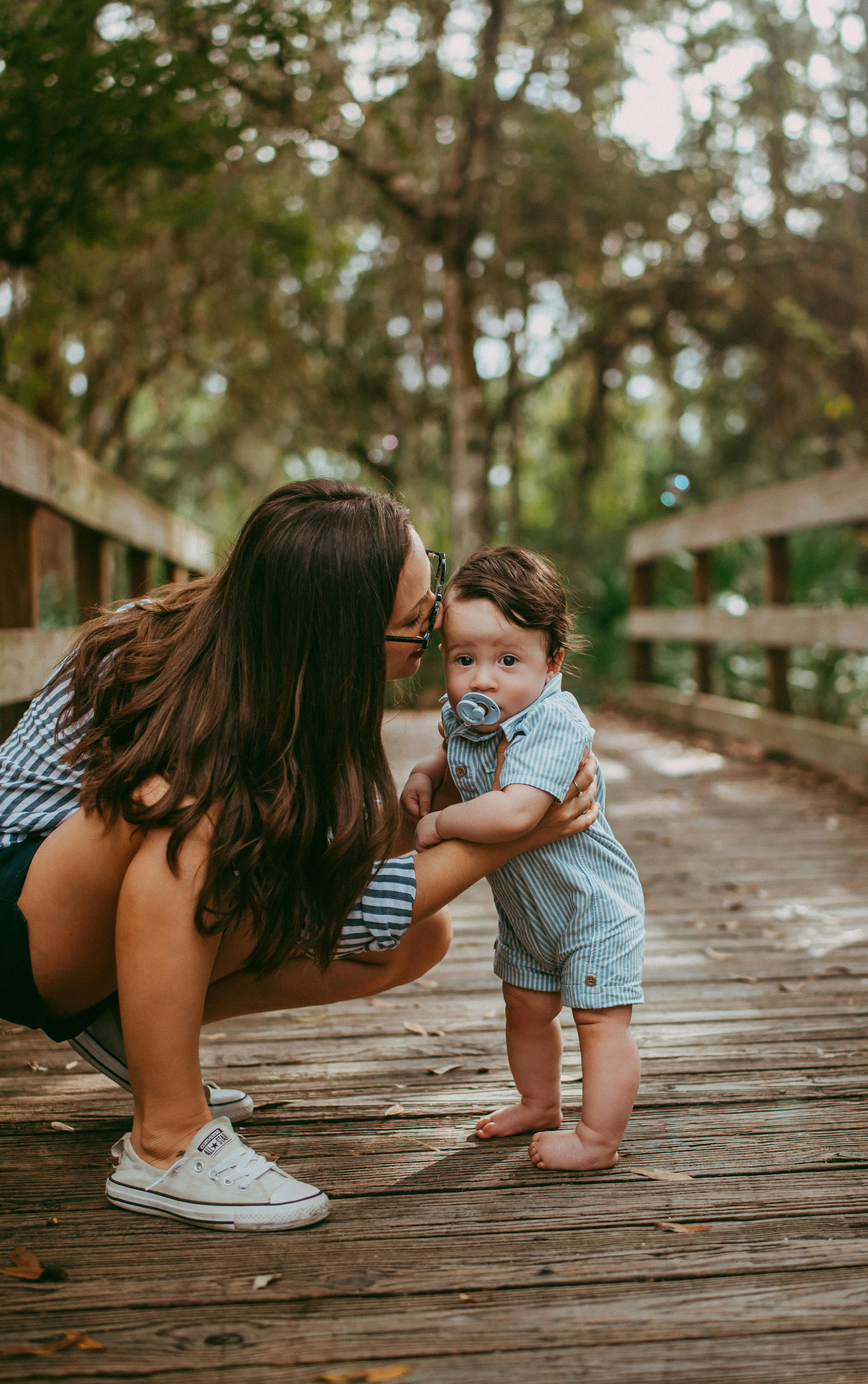 Side view young caring mother in casual summer clothes hunkering down and kissing adorable barefoot little son while standing on wooden footbridge in lush park