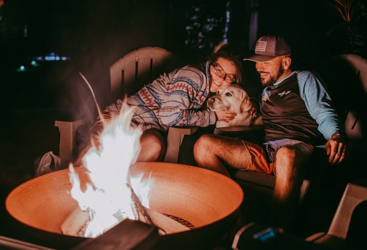 Cheerful Young Couple With Dog Recreating Near Campfire At Night