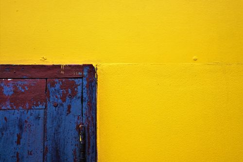 Free Close-up of a vibrant yellow wall paired with a rustic, textured blue door, showcasing vivid contrast. Stock Photo