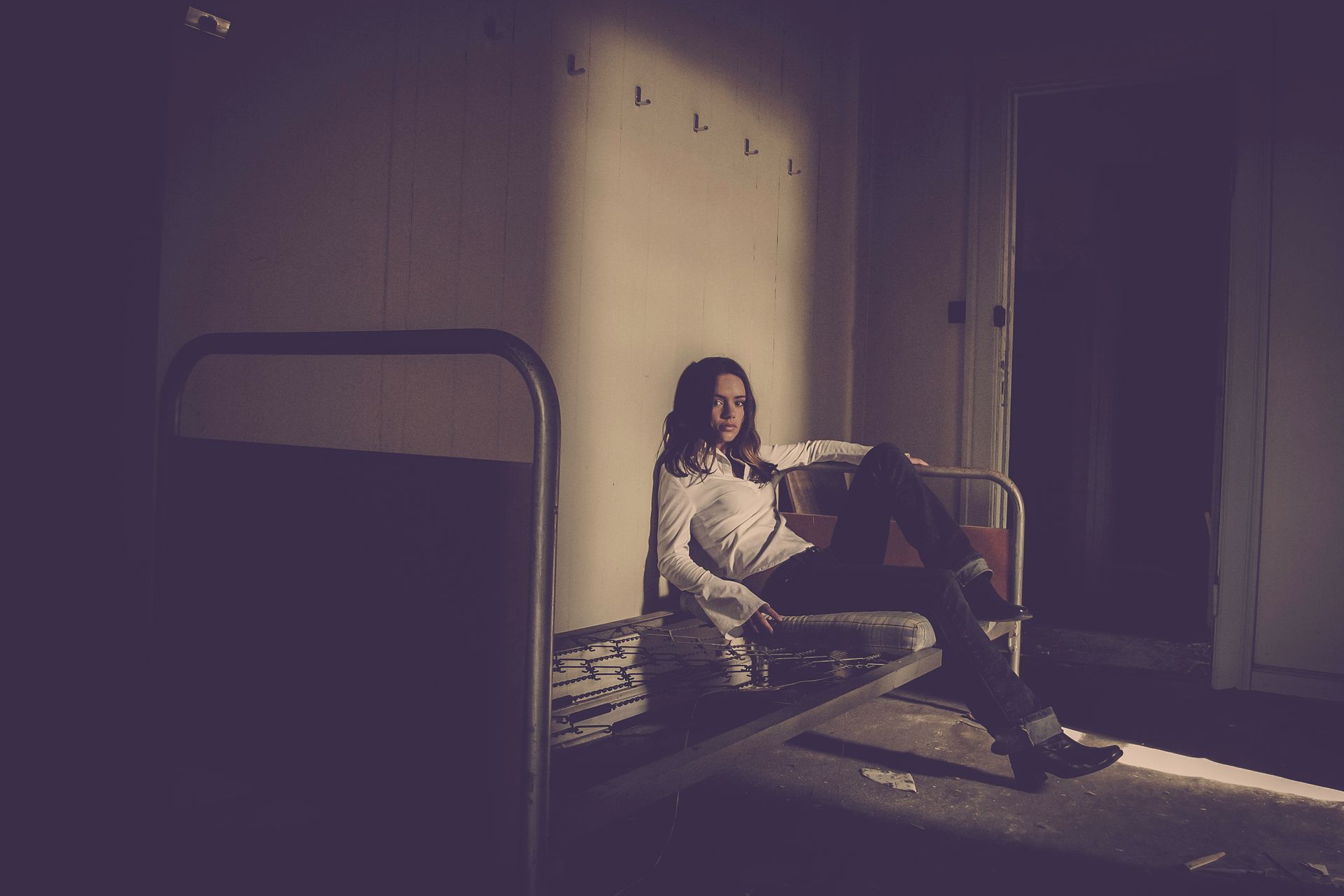 A woman leans on a vintage bed frame, in a dimly lit room, evoking a sense of mystery and introspection.