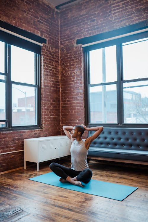 Full body of concentrated African American female stretching body in Sukhasana with hands behind head during yoga practice