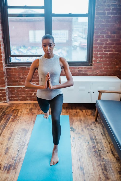 Concentrated barefoot African American female standing in High Lunge posture with namaste gesture while training yoga