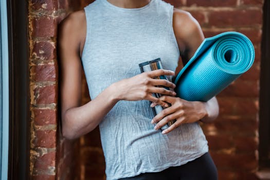 A fit young woman holding a yoga mat and water bottle, standing by a brick wall indoors.