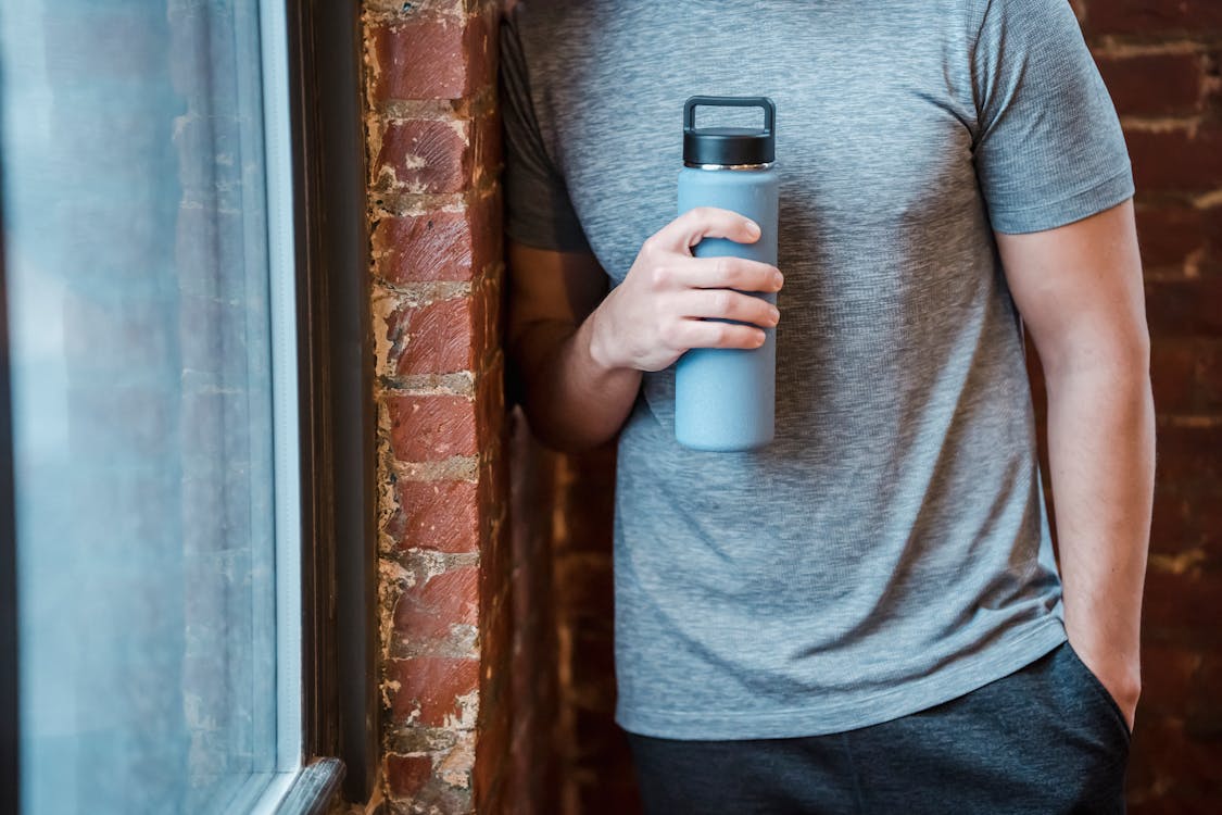 Unrecognizable muscular male in sportswear standing with hand in pocket near brick wall and window with bottle in hand in gym