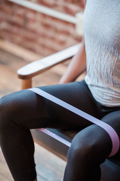 Unrecognizable sportive strong female wearing black leggings exercising with fitness expander on legs while sitting on couch on blurred background