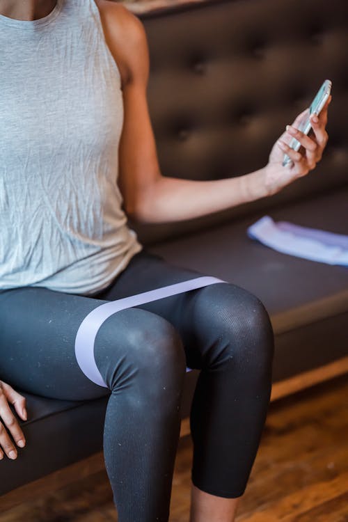 Free Faceless sportswoman in activewear sitting on couch and surfing internet on smartphone while exercising with elastic resistance rope on legs during training Stock Photo