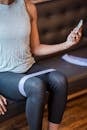 Faceless sportswoman in activewear sitting on couch and surfing internet on smartphone while exercising with elastic resistance rope on legs during training