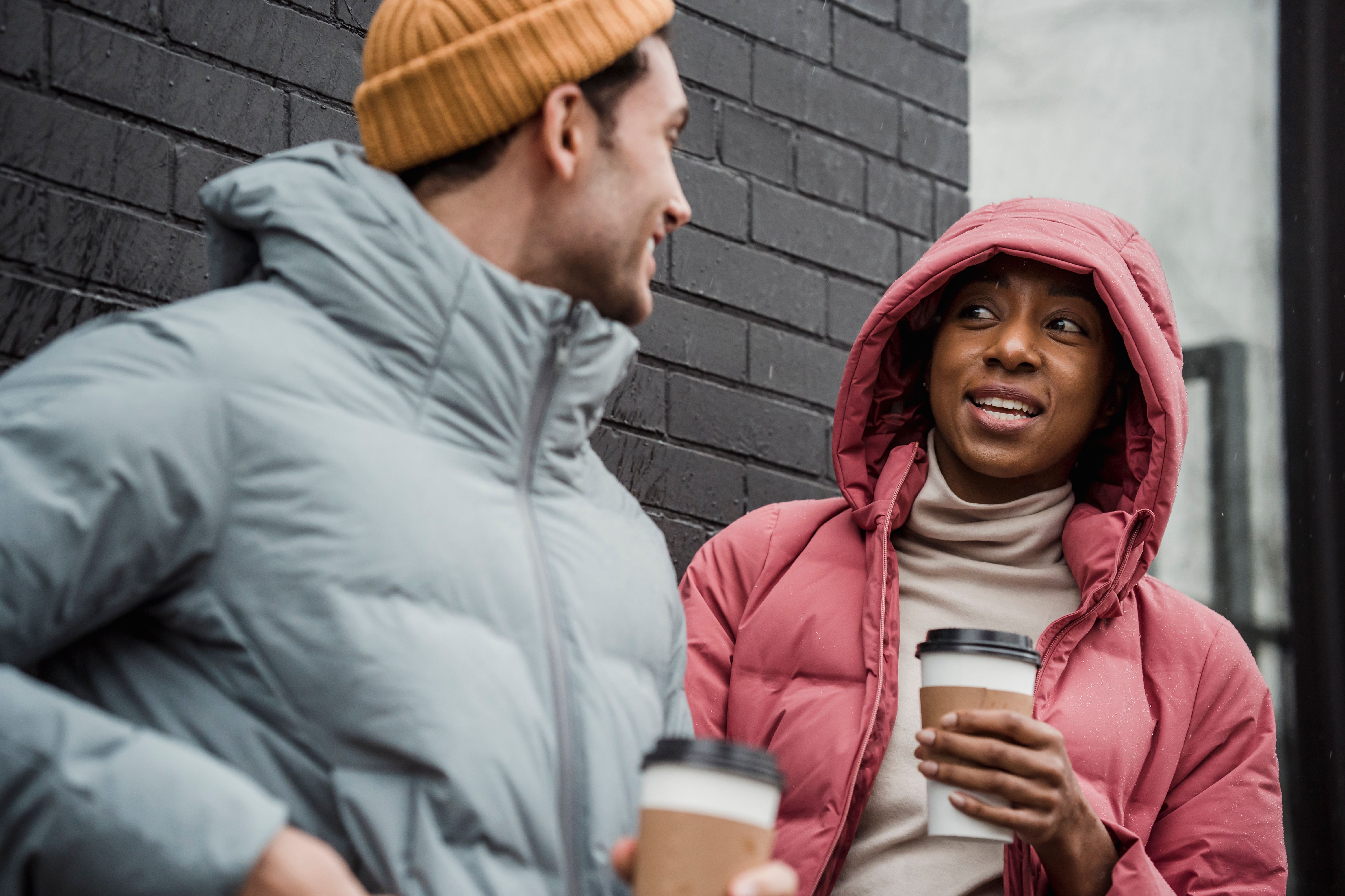 Cheerful multiracial couple in warm outerwear with takeaway hot beverages looking at each other while standing on street in overcast weather
