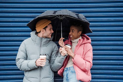 Positive multiracial couple in warm outerwear with takeaway coffee looking at each other while standing on street in rainy weather