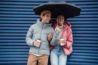 Happy multiracial couple with umbrella in warm outerwear laughing while standing on street with takeaway beverages in cold rainy weather