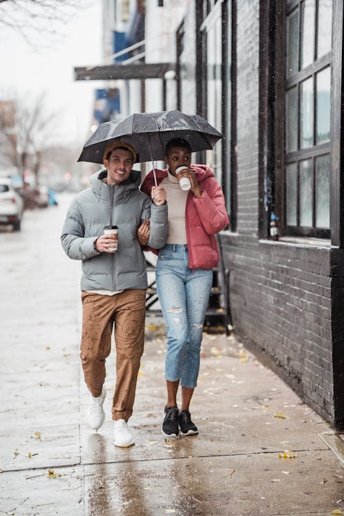 Cheerful diverse couple walking on street with coffee on rainy day