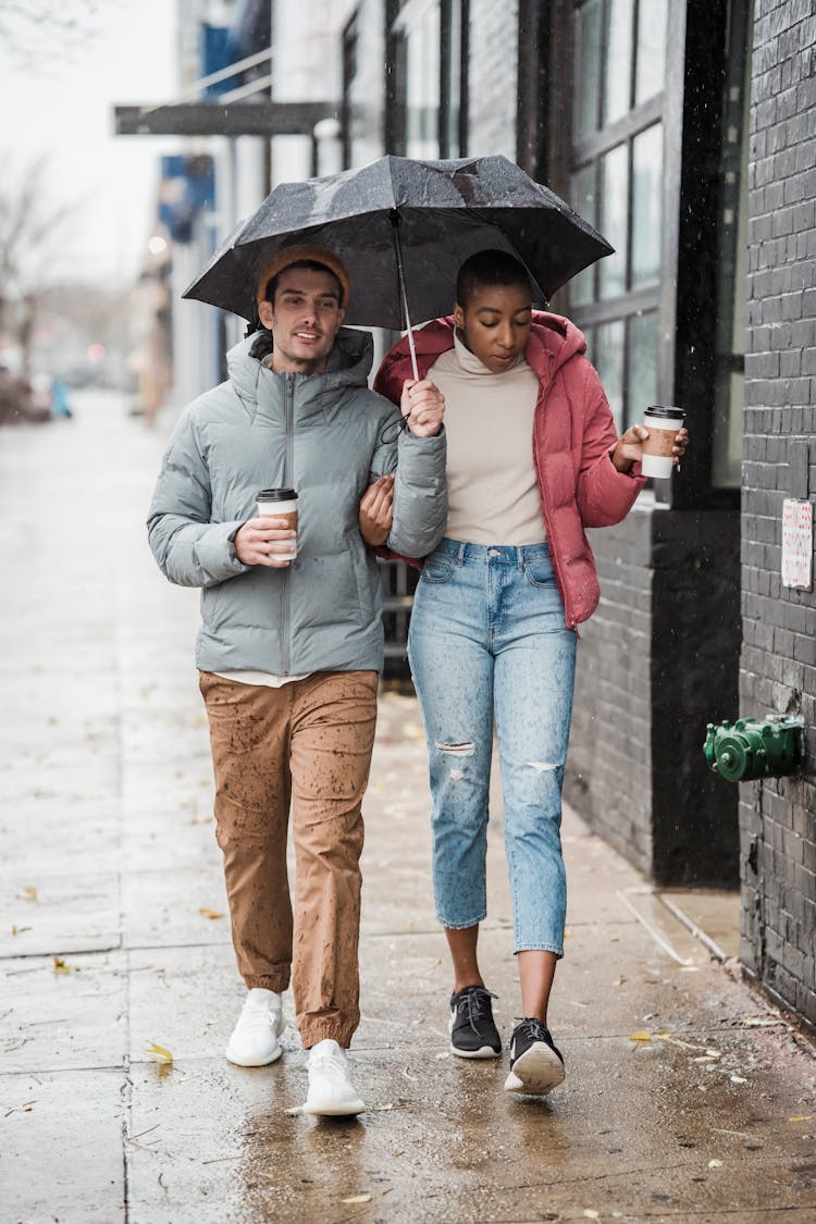 Content Diverse Couple With Umbrella And Coffee On Street