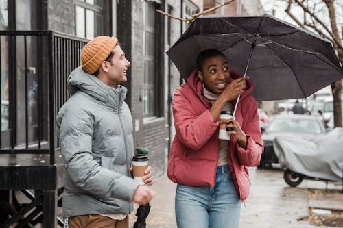 Free Optimistic diverse couple with takeaway hot drinks standing on wet sidewalk with umbrella on rainy and windy weather in city Stock Photo