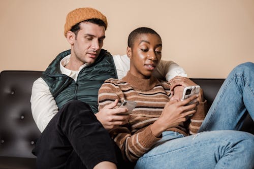 Free Concentrated multiracial loving couple surfing internet on cellphones while hugging on sofa in living room while spending time together at home Stock Photo