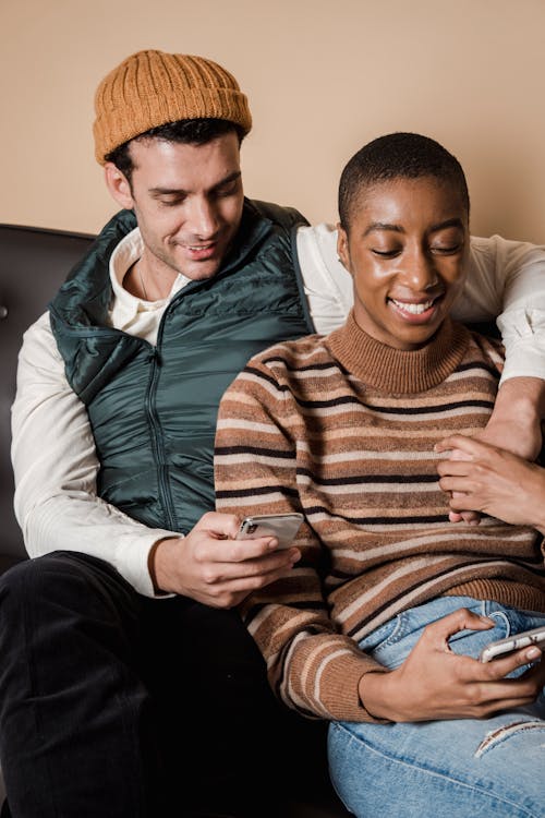 Free High angle of positive multiethnic couple holding hands while browsing mobile phones sitting on sofa Stock Photo