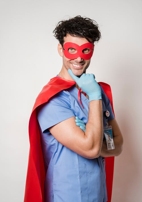 Positive male doctor in superhero mask and cape touching chin and looking at camera representing concept of COVID protection