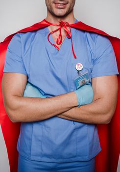 Positive male practitioner with folded hands wearing uniform and gloves with superhero cape for protection of coronavirus