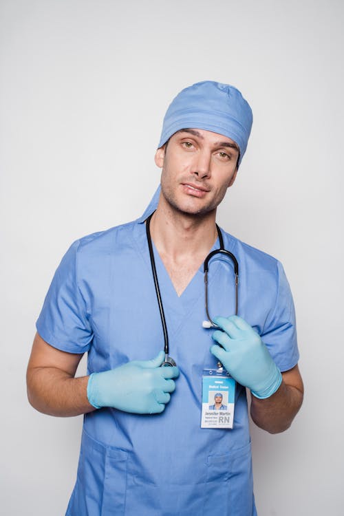 Free Pensive man doctor in blue uniform and sterile gloves standing with stethoscope and looking at camera against light background Stock Photo