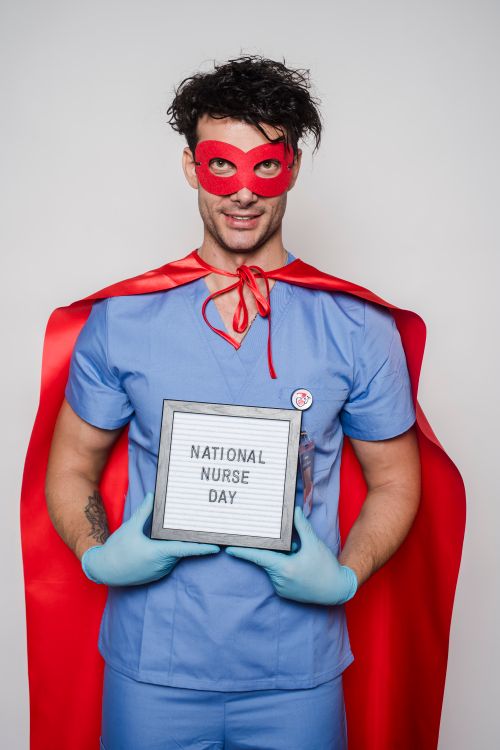 Free Positive man doctor in superhero costume and sterile gloves demonstrating card with National nurse day inscription against light wall in room Stock Photo